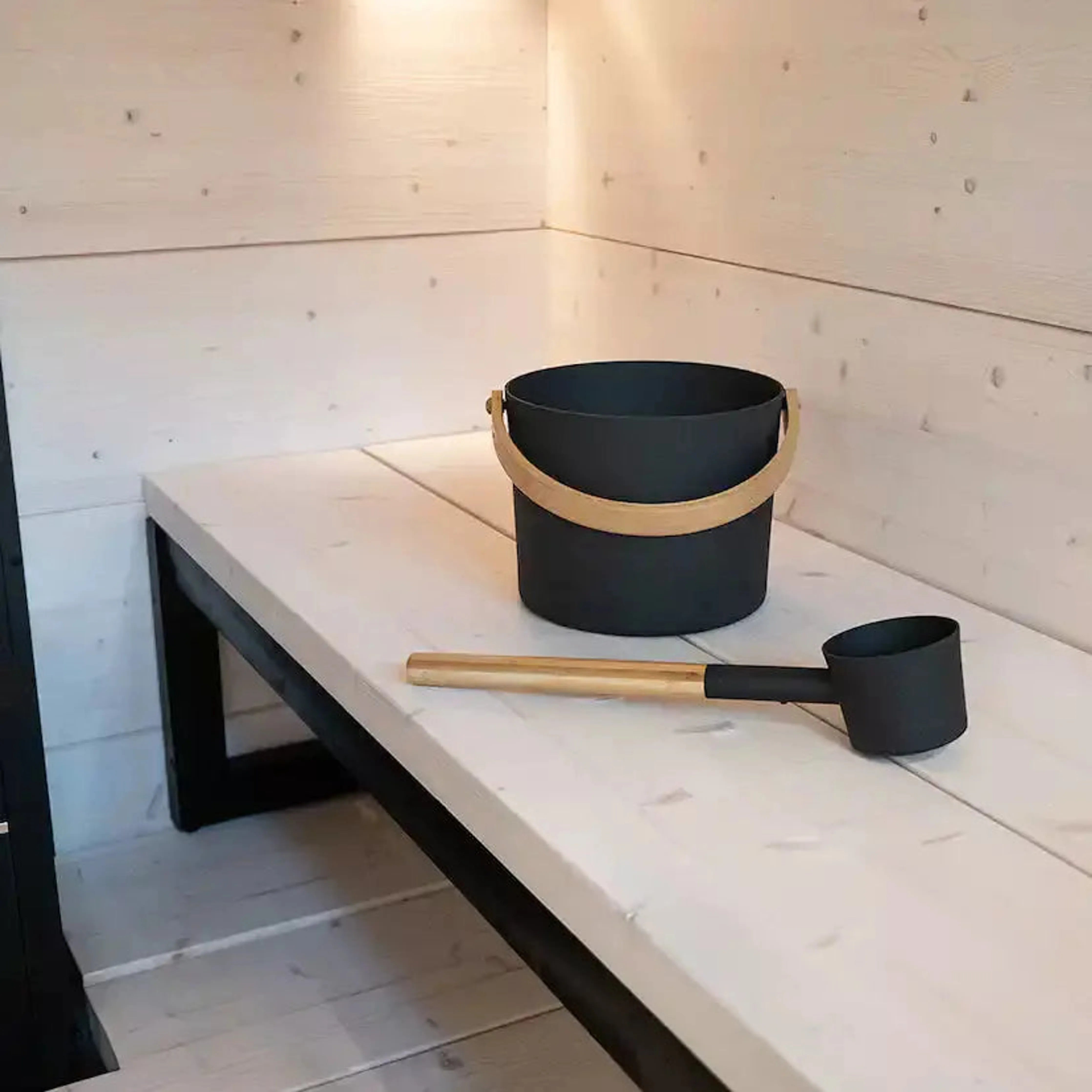 Harvia Black Sauna Pail and Ladle Set, black pail and wooden handled ladle on a light wood bench inside a sauna