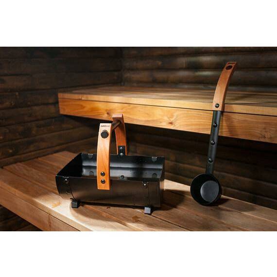 Harvia Legend Ladle SASPO101 Sauna Ladle, hanging on a wall hook beside a wood handled ember carrier in a wood paneled sauna interior.