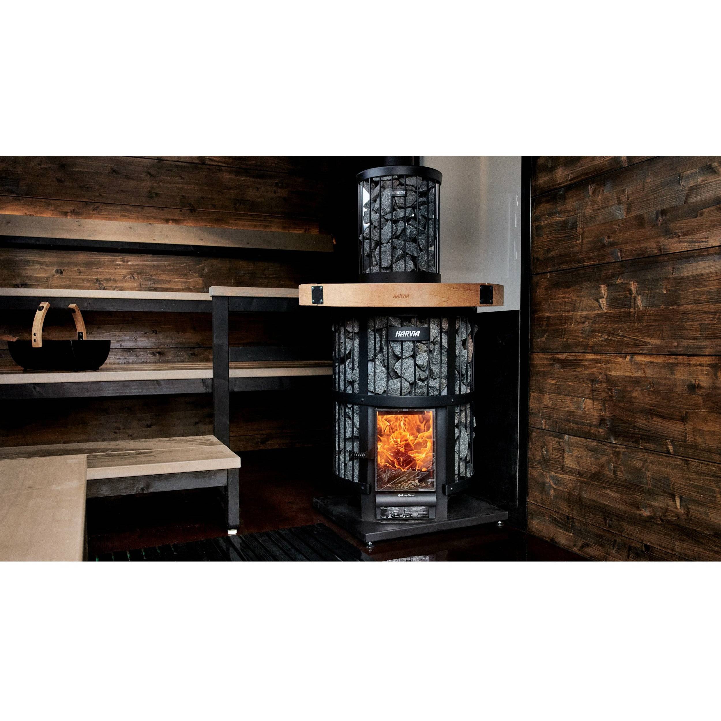 Harvia Legend 240 GreenFlame 21kW Wood-Burning Sauna Stove installed in a wood paneled sauna corner with a lit fire and a log basket nearby.