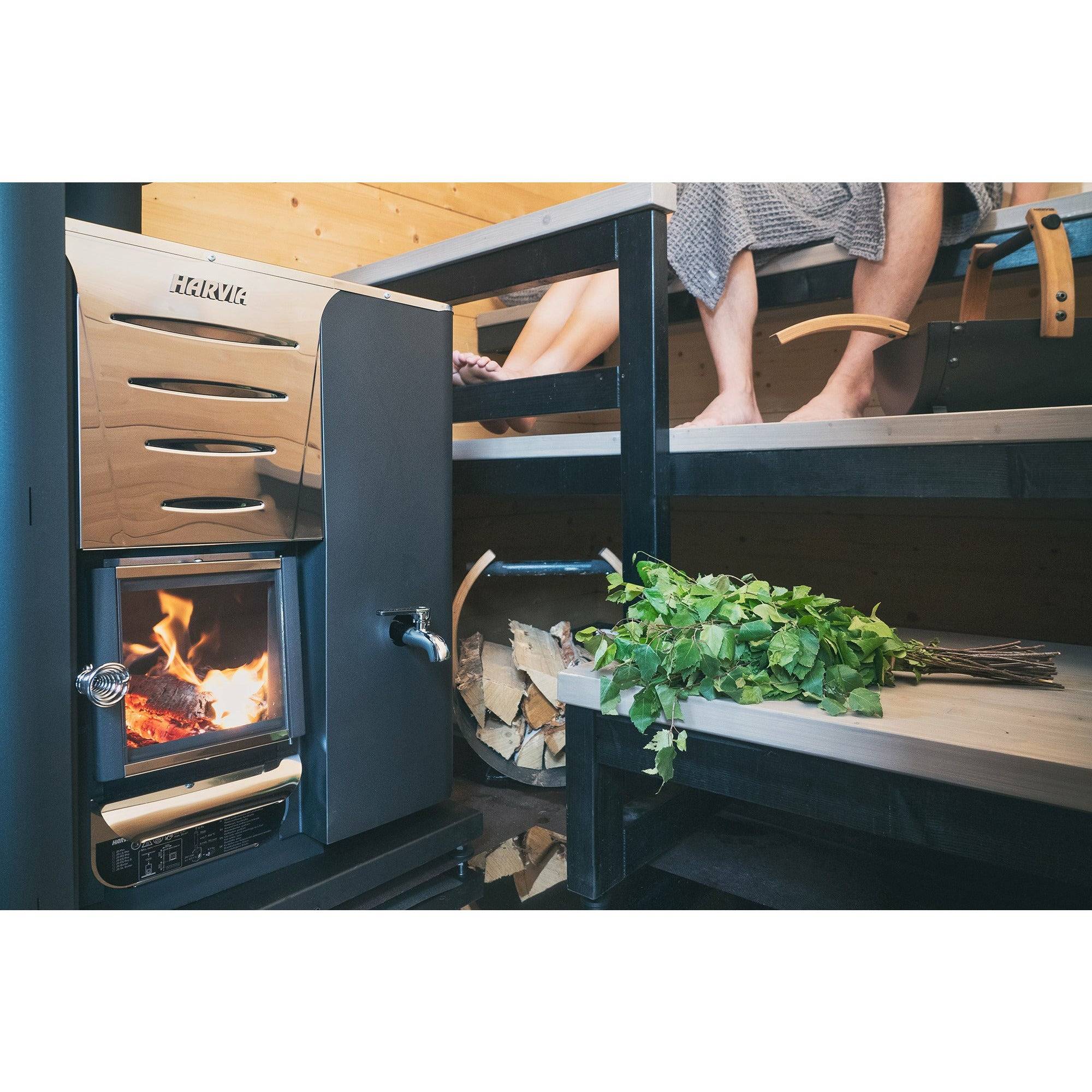 Harvia Pro 20 LS Wood-Burning Sauna Stove with Water Tank, installed in a wooden sauna with fire burning, firewood and a birch whisk nearby.