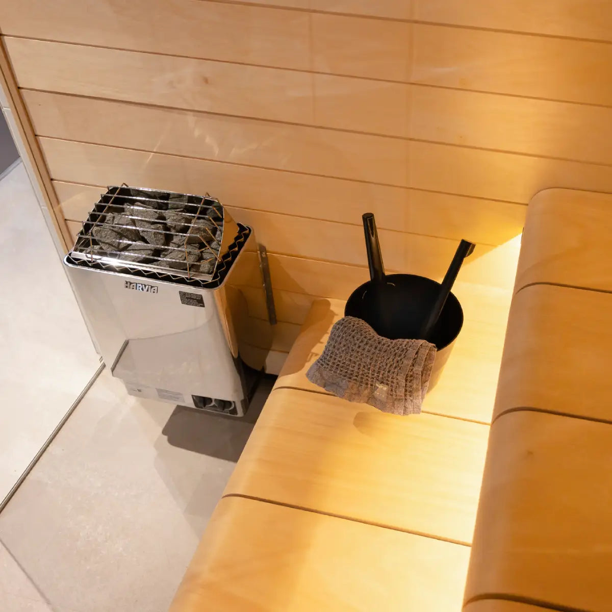 Harvia KIP60W 6kW Electric Sauna Heater mounted in a sauna corner beside a bench with a black bucket, ladle, and towel
