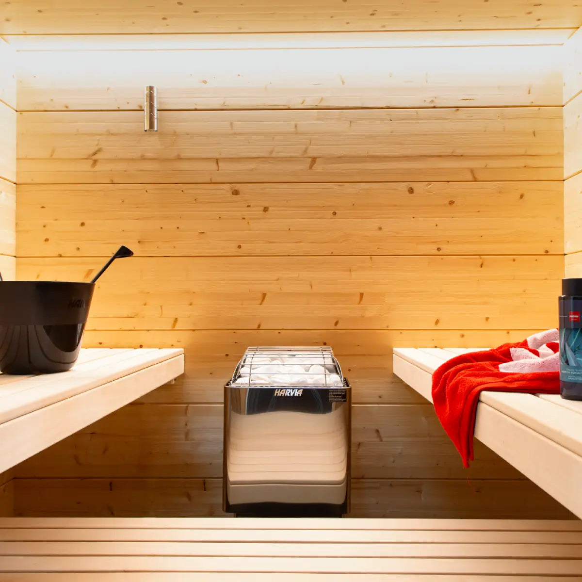Harvia KIP80W 8kW Electric Sauna Heater installed in a wood paneled sauna, flanked by benches with a sauna bucket and red towel.