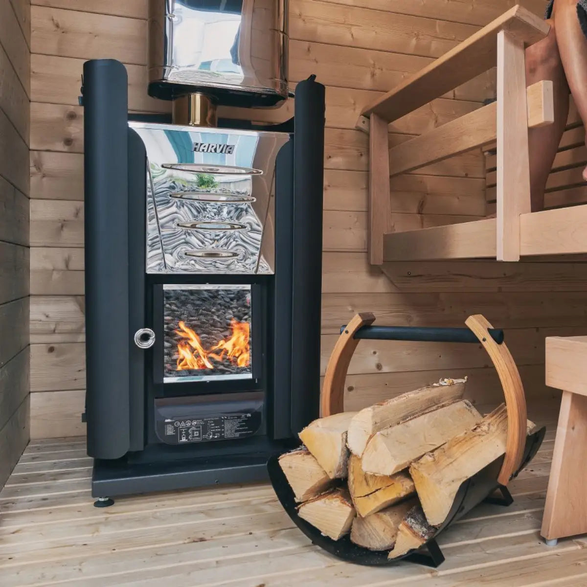 Harvia Pro 20 24kW Wood-Burning Sauna Stove with mirrored steel top and lit firebox, log basket and stacked firewood beside it.