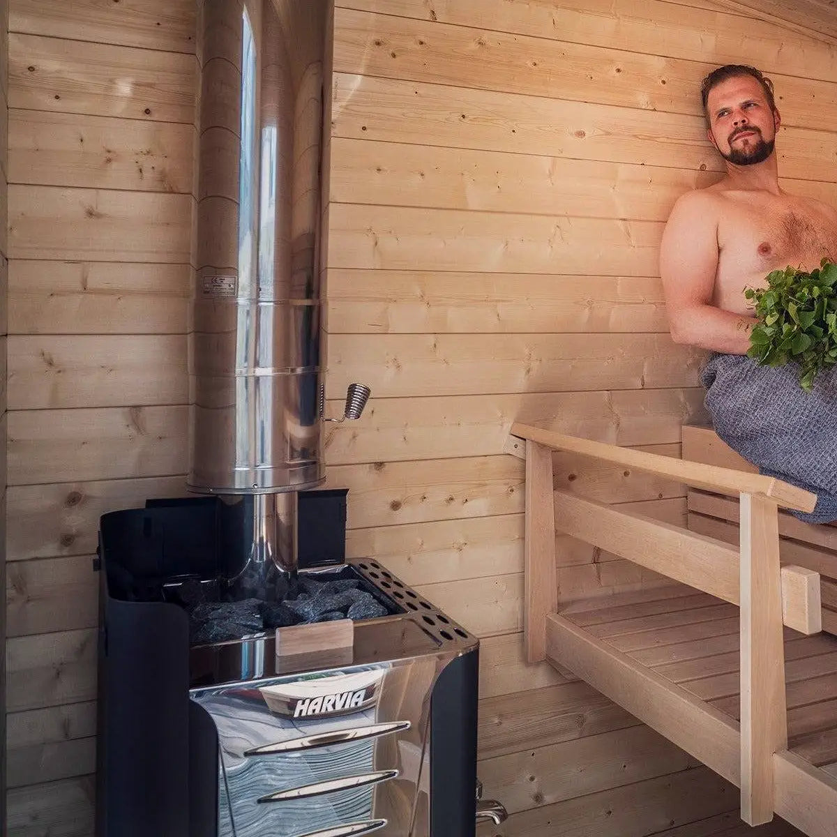 Harvia Pro 26 26.6kW Wood-Burning Sauna Stove installed in a wood sauna, man on bench holding a vihta whisk beside the chimney