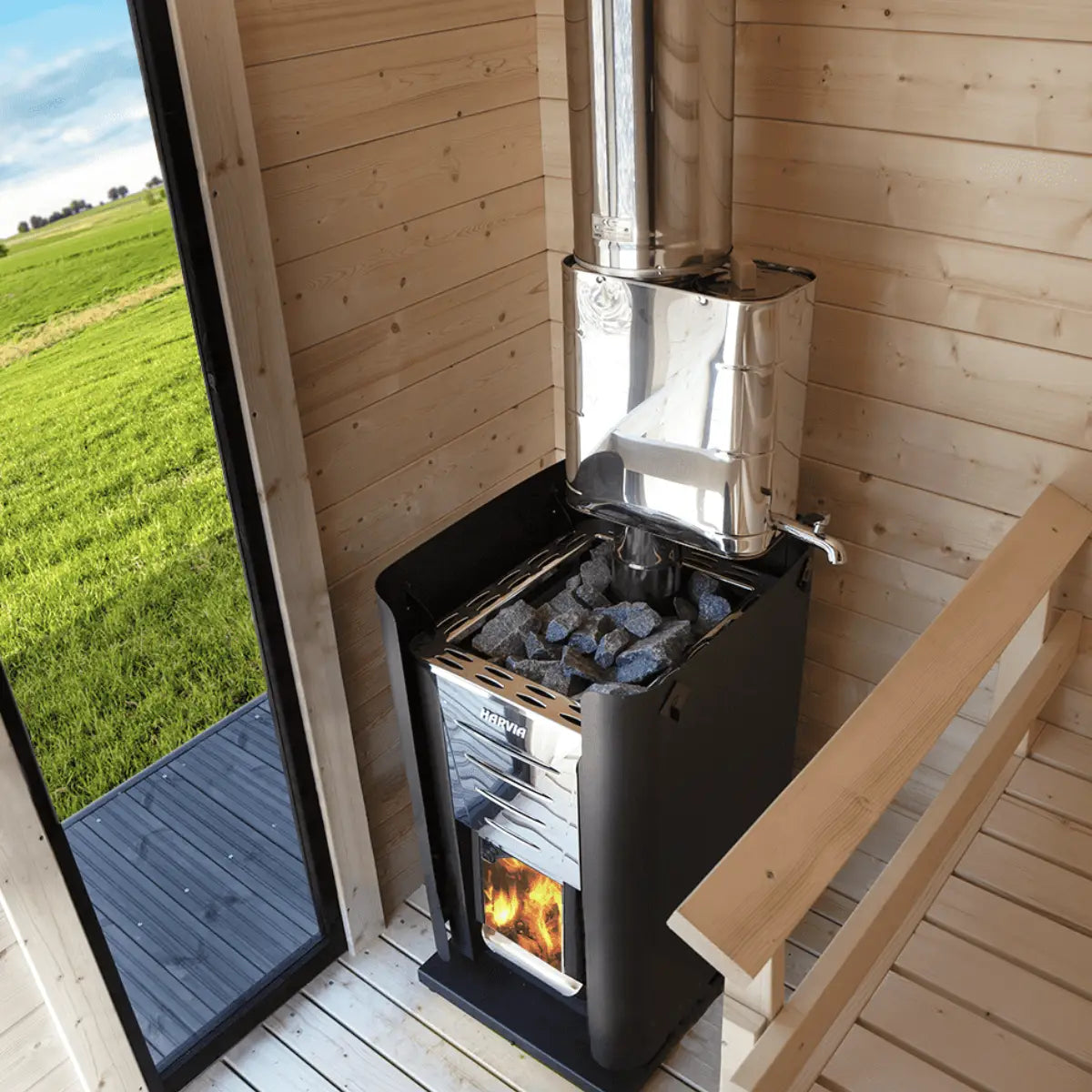 Harvia Pro 26 26.6kW Wood-Burning Sauna Stove in a cabin corner with chimney pipe, stones, and lit fire, open door to a green field