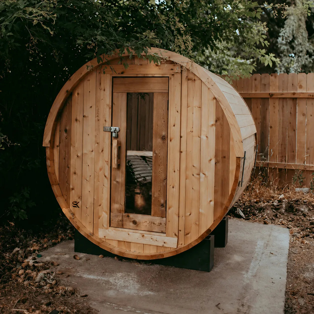 The Sweat Barrel cedar barrel sauna installed on a concrete pad in a backyard with cedar fence and trees, front view through glass door.