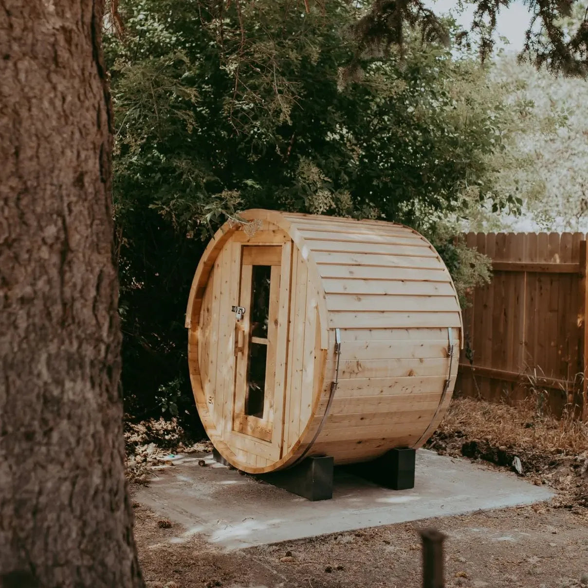 The Sweat Barrel cedar sauna installed under shade trees in a private backyard on a concrete pad with wood fence.