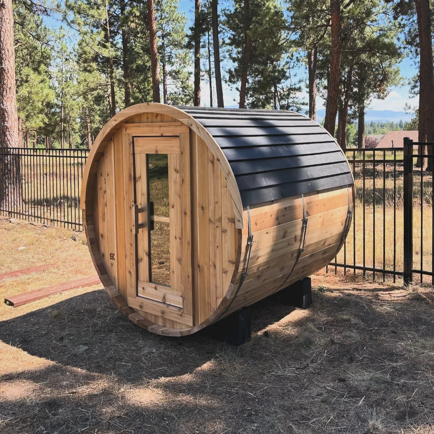 Real customer install of The Sweat Barrel cedar sauna with metal roof in a forested mountain yard with pine trees and distant mountains.