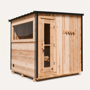 The Summit cedar 4-6 person outdoor sauna three-quarter angle on white background showing slanted black-trim metal roof, glass door, side window, and exterior cedar peg rack.