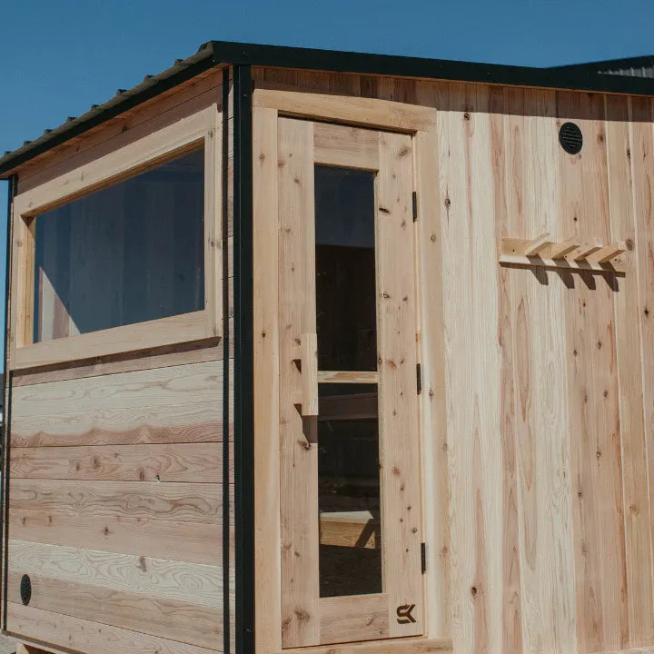 Close-up of The Summit cedar sauna exterior showing the slanted black-trim roof, side picture window, glass door, and exterior cedar peg rack.