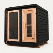 The Sweat Cabin Blackout Edition charred cedar 4-person sauna three-quarter angle on white background showing rounded corners, glass side panel with heater visible, and natural cedar door frame.