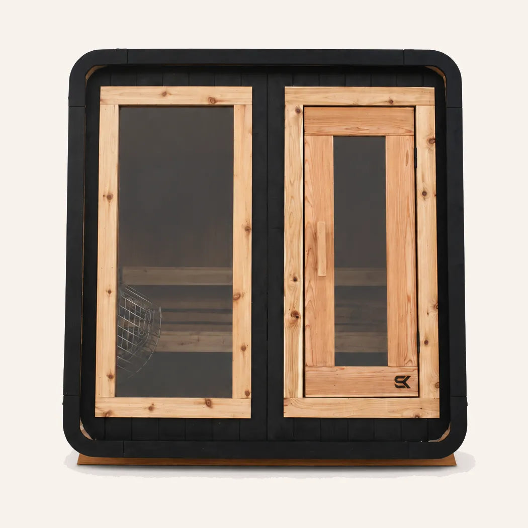 The Sweat Cabin Blackout Edition charred cedar 4-person sauna front view on white background showing contrasting natural cedar door, side glass panel with stone basket heater, and rounded black frame.