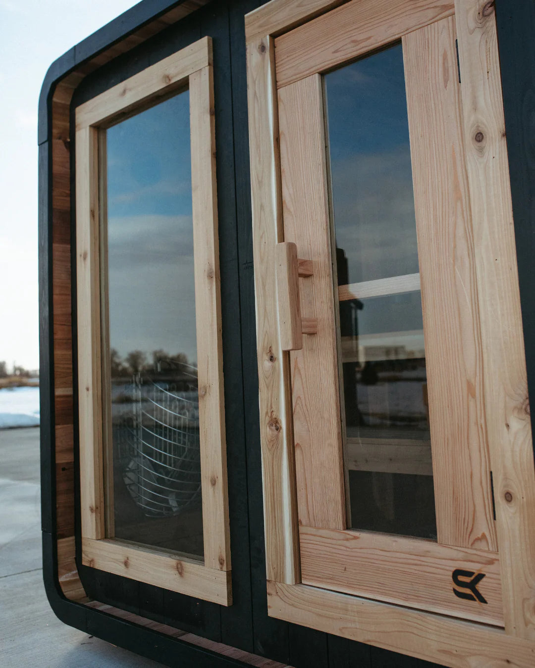Close-up of The Sweat Cabin Blackout Edition showing the natural cedar door frame, glass panel with stone basket heater visible, and Sweat Kingdom SK logo on charred cedar siding.
