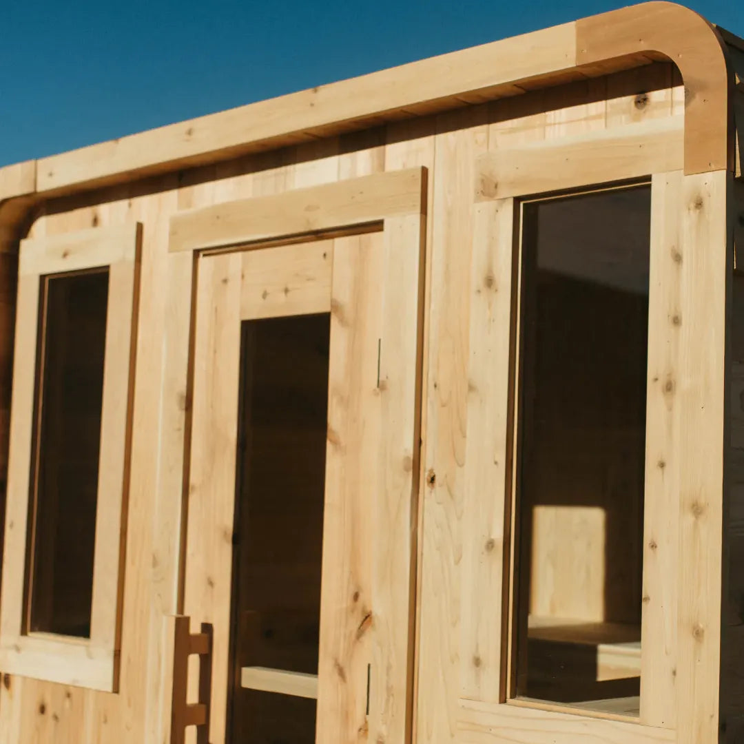 Close-up of The Sweat Cabin Deluxe cedar 6-person sauna exterior showing the central glass door, two side windows, and rounded top corner against a blue sky.