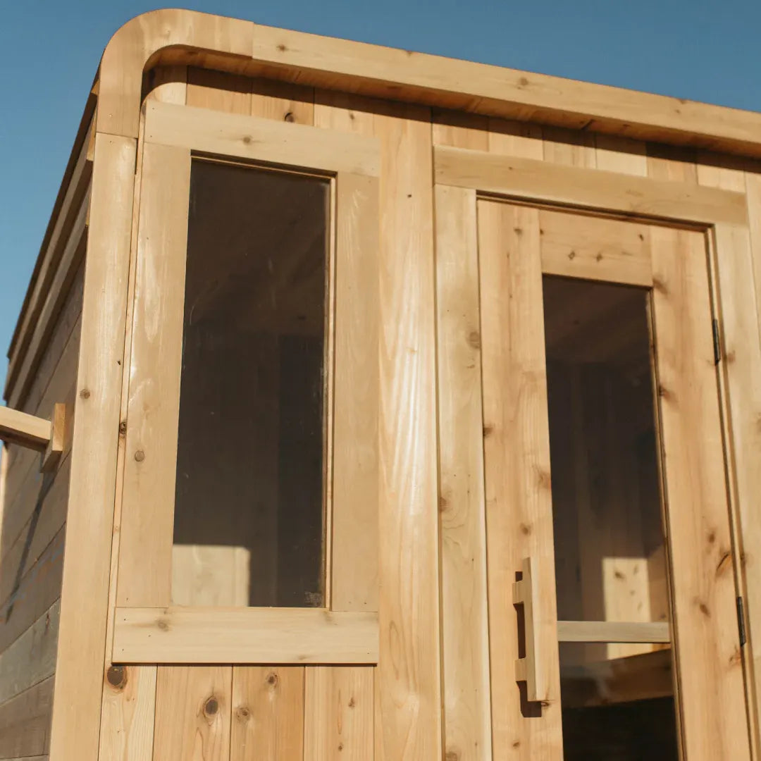 Close-up of The Sweat Cabin Deluxe showing the side picture window beside the open glass door with cedar door handle detail.