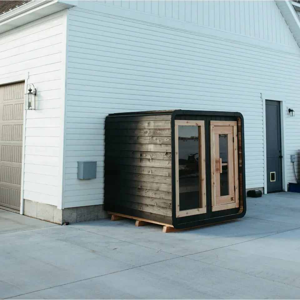 The Sweat Cabin Blackout Edition charred cedar sauna installed beside a white-sided manufacturing building with concrete pad and overhead light.