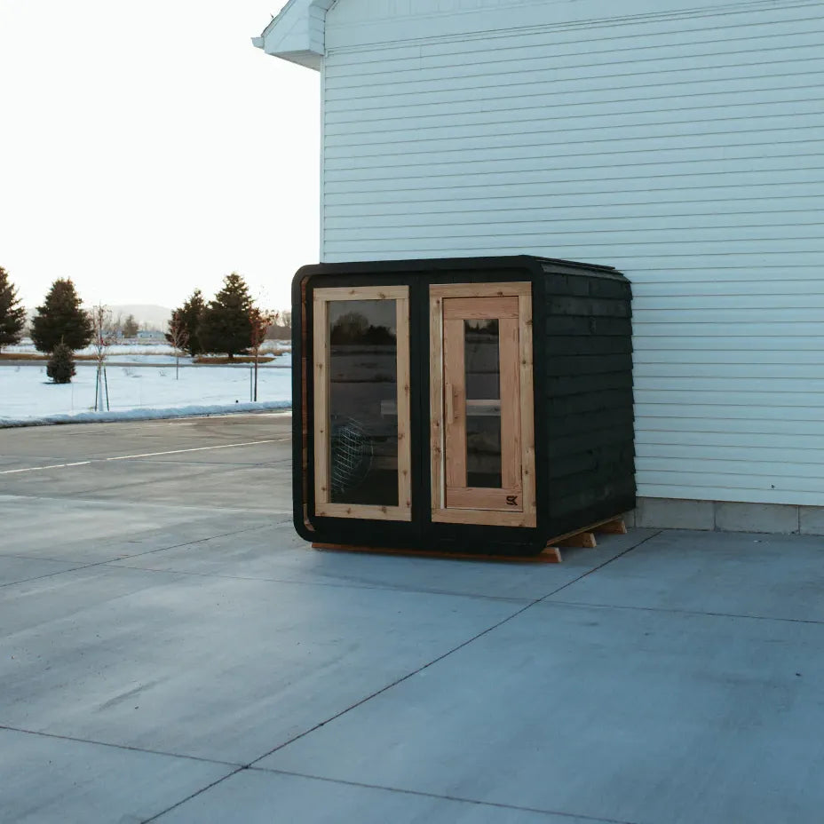 The Sweat Cabin Blackout Edition charred cedar sauna shown outside against a white building with snow-covered field and pine trees at sunset.