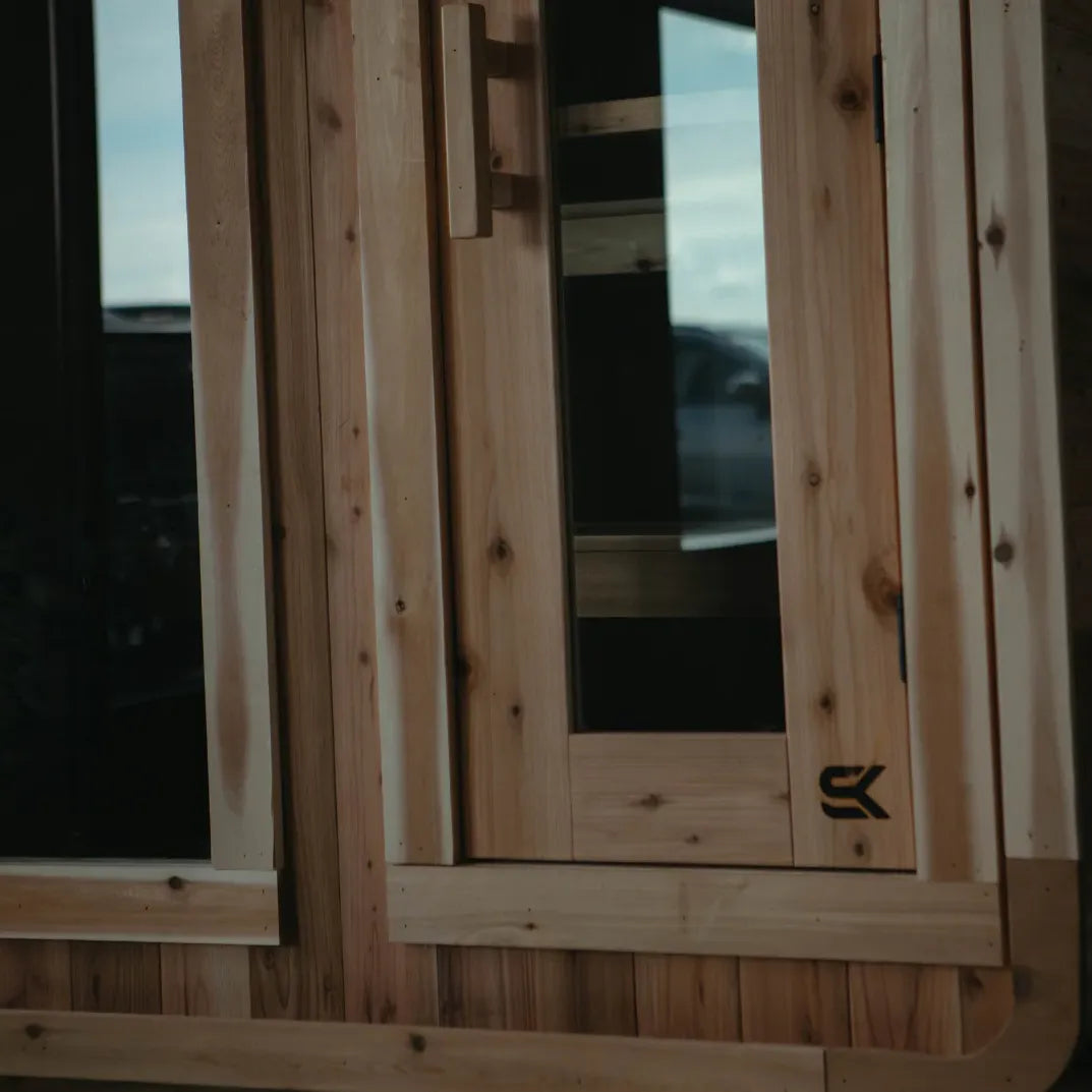Close-up detail of the cedar door and large picture window on The Sweat Cabin sauna with Sweat Kingdom SK logo.