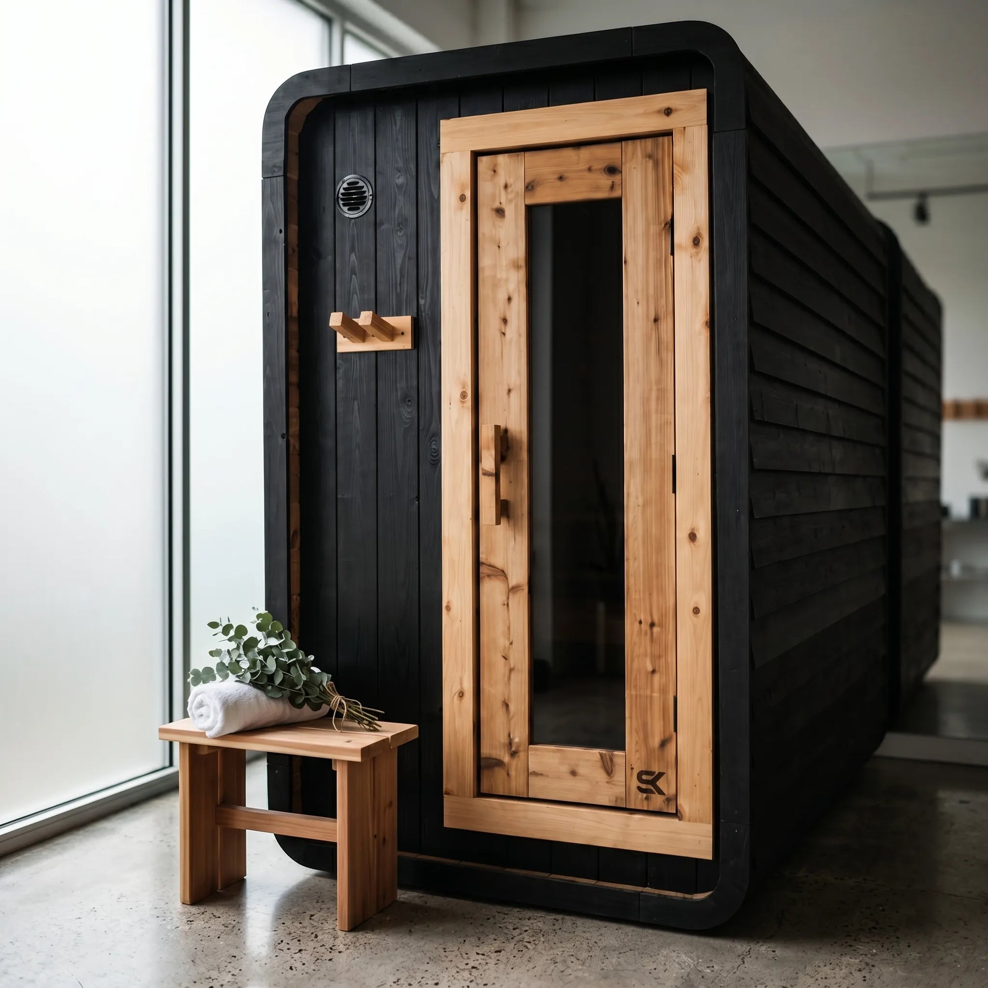 The Sweat Pod Black Edition charred cedar sauna in a modern minimalist studio with cedar bench, eucalyptus, and concrete floor.