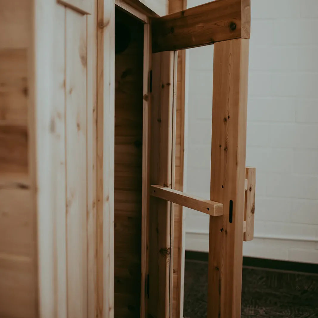 The Sweat Pod cedar sauna door open showing the cedar interior and light tile bathroom wall behind.