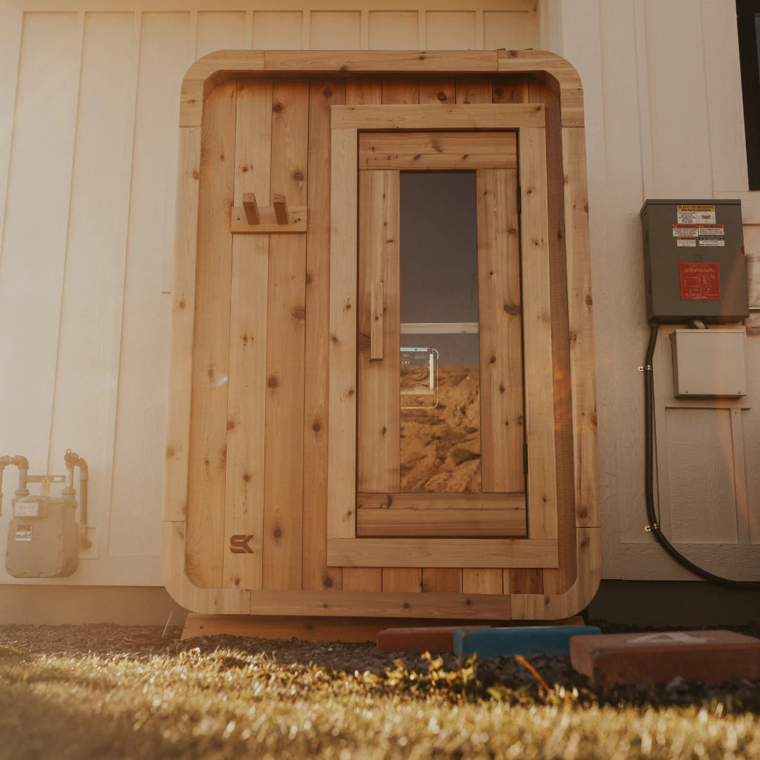 The Sweat Pod cedar indoor sauna installed beside a residential home at golden hour, front view with glass door.