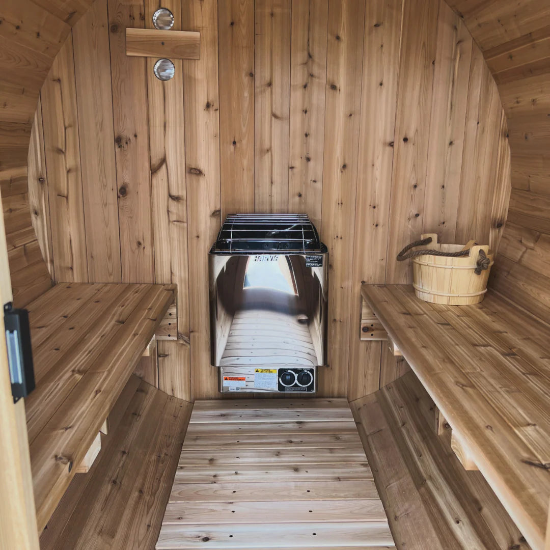 Inside The Sweat Barrel sauna showing Harvia electric heater, two-tier cedar benches, and traditional wooden sauna bucket.