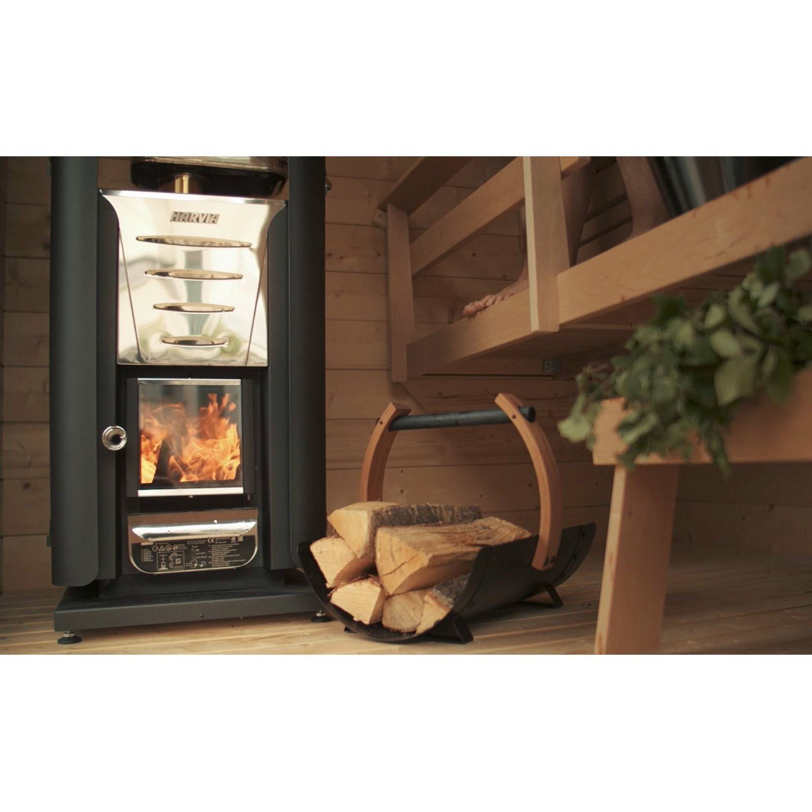 Harvia Legend Firewood Basket SASPO102 filled with logs beside a lit wood burning sauna heater inside a wood paneled sauna room