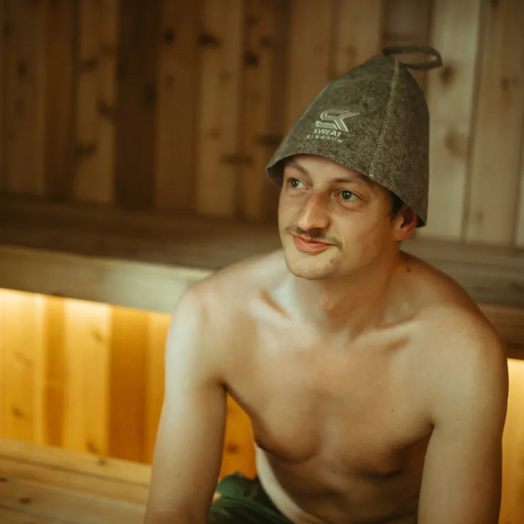 Man wearing a gray Sweat Kingdom sauna hat while sitting inside a wooden sauna